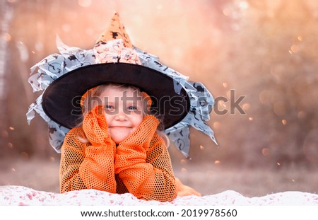 Happy Halloween concept.Cute funny little child in magical wizard hat lies on blanket and propping head in her hands on festive background. Smiling baby girl in witch orange costume with long sleeves 