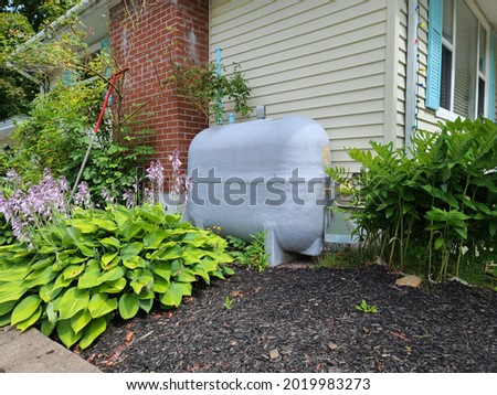 An oil tank hidden on the side of a suburban house in a close knit community. The tank is painted gray and nestled next to a brick chimney and flower beds filled with Hosta plants. Royalty-Free Stock Photo #2019983273
