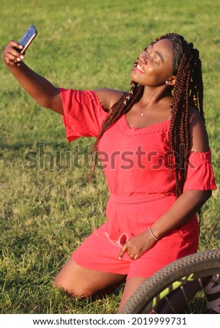 Young African girl in a dress on the nature. A black woman from Cameroon takes a picnic in the summer. The girl takes pictures of herself using the phone.