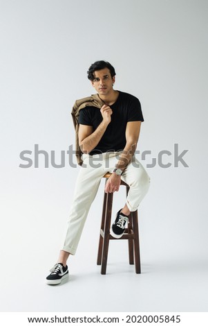 full length of young man holding blazer while sitting on chair on grey Royalty-Free Stock Photo #2020005845
