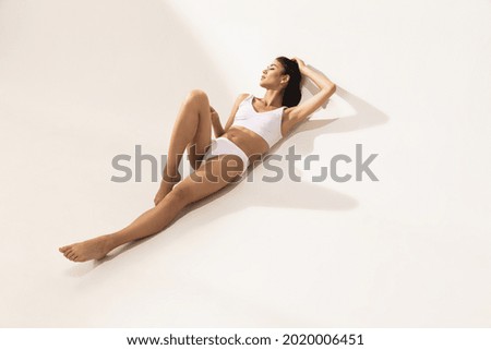 One young beautiful tanned woman with perfect figure in white underwear lying on floor over white studio background. Copypace for ad. Concept of female beauty, tenderness, body and skin care