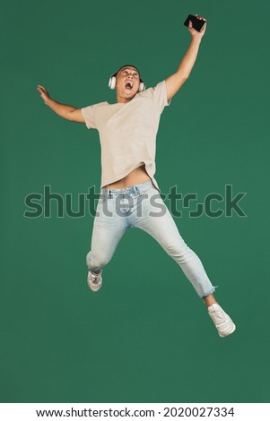 Enjoying music. Portrait of young handsome asian man, guy in headphones jumping with smartphone isolated over dark green studio background. Concept of human emotions, facial expression, youth.