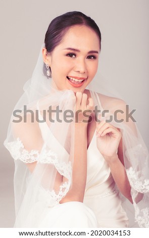 Portrait of young attractive Asian woman wearing white wedding gown holding her veil smiling at camera. Concept for bride pre wedding photography.