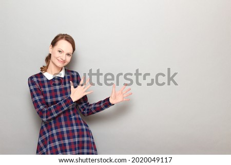 Studio portrait of happy joyful girl wearing checkered dress, smiling, raising hands up, dancing, celebrating victory or success, being in good mood, standing over gray background, with copy space