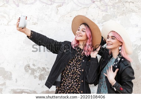 Two happy beautiful young stylish girls with pink hair in fashionable denim clothes with straw hats take a selfie photo near a white wall