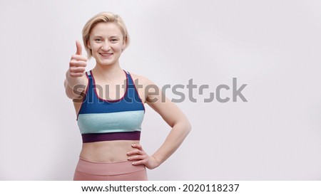 Girl dressed in activewear making thumbs-up sign at the camera. Strong girl posing for the camera showing her strength. Concept of fitness and gym. Isolated over white background studio.