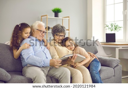 Happy grandparents with twin grandchildren browse family photo album and share happy memories. Family having fun at home sitting on sofa in living room. Concept of family history and memories.