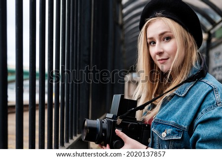 Young woman taking pictures with a medium format film camera. She is wearing a denim jacket and a black beret. Looking to camera.