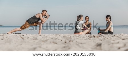 Panoramic shot of photographer taking picture group of woman getting rest after fitness exercises on the beach. Vlogging, blogging concept. Photoshot of young athletes, sportswomen outdoors.