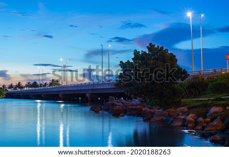 Beautiful sunset and sunrise on the beach in the summertime surrounded by rocks, trees, palms, a bridge, some mountains in the horizon and a calm water reflecting clouds and lights.