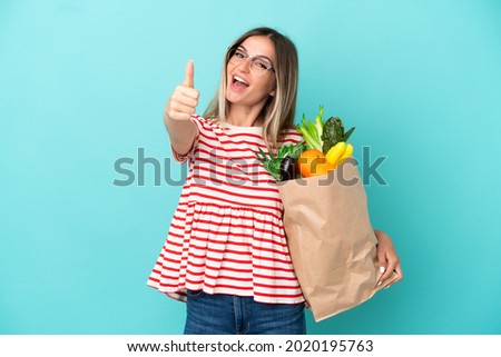 Young woman holding a grocery shopping bag isolated on blue background with thumbs up because something good has happened