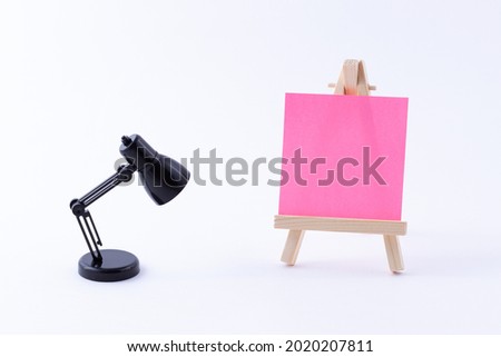 Wooden Easel Miniature with Blank Colored Square Canvas or Memo Paper - Mockup. Mini Wooden Stand with Clean Artboard and Small Black Table Lamp on White Background, Copy Space