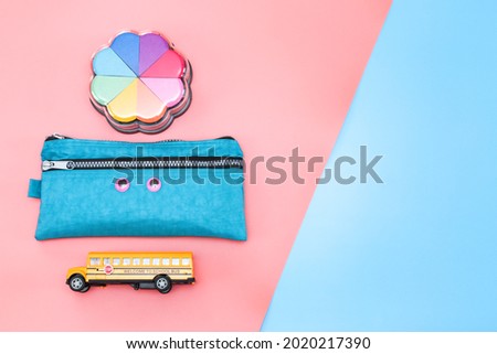 A set of accessories from: school bus, pencil case and paint with rainbow sponges lie on the left on a pink and blue background with space for text on the right, top view close-up. Concept back to sch
