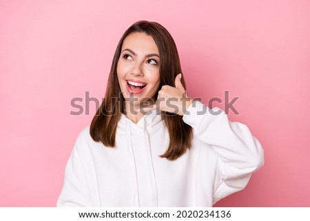 Photo of impressed cute young lady wear white hoodie smiling showing phone gesture looking empty space isolated pink color background