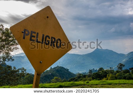 a yellow danger traffic sign in front of a beautiful valley of green mountains and a spectacular cloudy sky