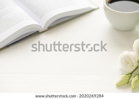 Photo of white cup of coffee flowers and open book on white table with copyspace