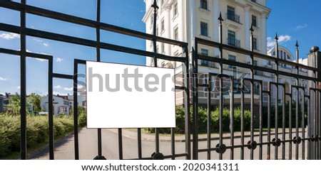 Blank white information sign board hangs on black metal fence against luxury building