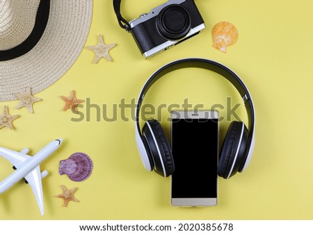 Top view of mobile phone with headphones around, camera, straw hat, airplane model sea shells and starfishes on yellow background. Summer beach music or podcast background.