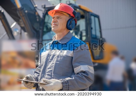 Industrial worker male senior in helmet holds tablet computer on background of production of escovator factory. Royalty-Free Stock Photo #2020394930