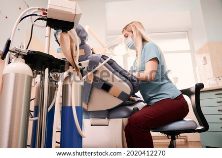 Side view of dentist treating kid teeth while child lying in dental chair. Equipment of inhalation sedation on foreground. Concept of children dentistry, stomatology and dental care. Royalty-Free Stock Photo #2020412270
