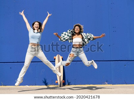 two multiethnic women jumping on a blue wall, with skateboard and casual clothes