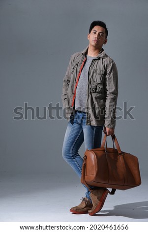 Full length portrait of a male in coat ,jeans holding handbag  standing isolated on gray background