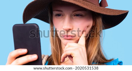 Close up photo of a pensive blonde girl with squints eyes wearing the brown hat looking at the phone holding in her hand. 