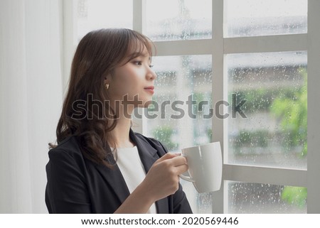 Beautiful Asian businesswoman (Thai, Japanese, Korean or Chinese) with a white coffee cup stands near the window on a rainy day happily and looks out the window. Royalty-Free Stock Photo #2020569446