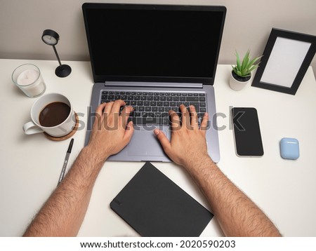Man's hands on a white table with a laptop typing on the keyboard next to a mobile phone a black notebook a cup of coffee a clock a white candle a small bush and a blank photo frame