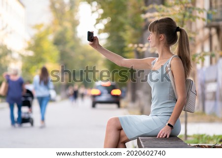 Young pretty woman taking selfie with mobile phone on warm summer day sitting on a city street bench outdoors.