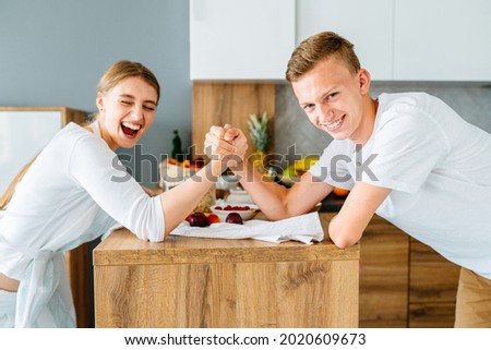 Teenage brother and her adult sister, young in 20s, arm wrestling at cozy home kitchen.