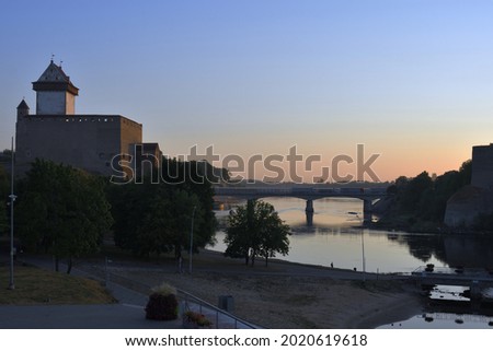 Narva Castle and the border river Narva with a bridge at dawn, the sun is not yet visible, but its rays are already painting the sky and the castle with the Long Herman tower in yellow-red colors.