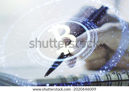 Double exposure of creative Bitcoin symbol hologram with woman hand writing in notepad on background. Mining and blockchain concept