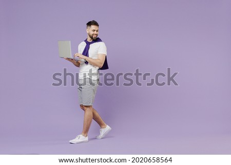 Full size body length side view young brunet man 20s wears white t-shirt purple shirt hold use work on laptop pc computer go move look back down isolated on pastel violet background studio portrait.