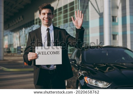 Bottom view young friendly traveler businessman man wear black suit stand outside at international airport terminal hold card sign with welcome title text waving hand Air flight business trip concept.
