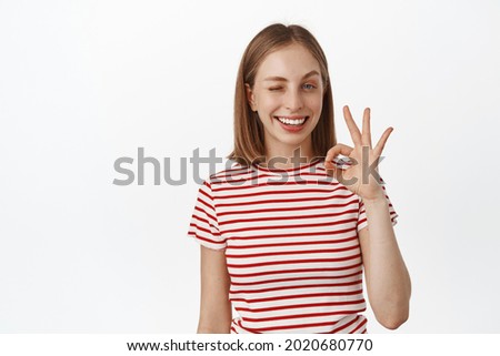 Beautiful blond girl smiling, winking and showing ok, okay Zero sign, assure everything good, all under control, no problem, praise and like smth, standing against white background