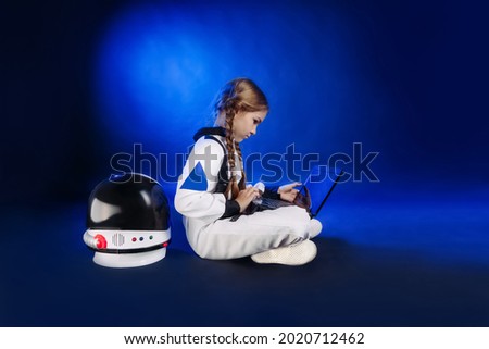 Teenager girl wearing astronaut costume using laptop sitting on dark blue background with copy space. Cute girl in spacesuit watching online distance learning lesson.