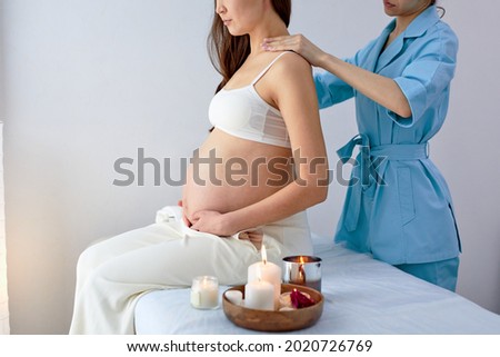 Masseur physiotherapist in blue uniform massaging back of pregnant woman in spa center, side view. Mom-to-be preparing to give birth in a short time. Pregnancy concept. Cropped photo
