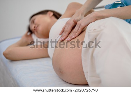 Pregnant young woman with beautiful skin lying on bed having relaxing prenatal massage, physiotherapist doing various techniques for better massaging, close-up photo of hands on tummy