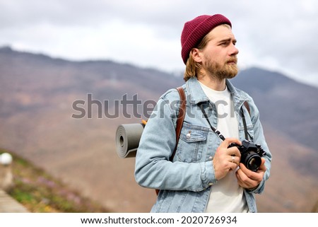 Traveler walking in mountains with camera, enjoying the scenery, side view portrait. Bearded caucasian male in casual outfit outdoors, with backpack. Copy space for advertisement