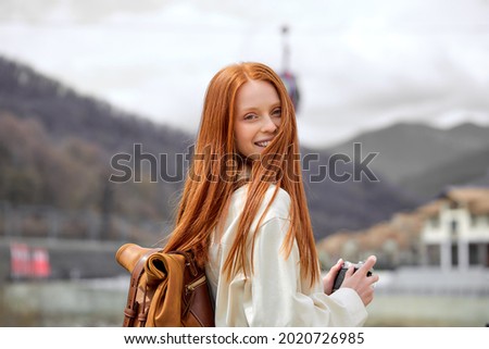 good-looking red-haired woman with retro camera in hands on trip, in mountains, in casual coat or jacket. traveling alone, spend holidays, take photos.