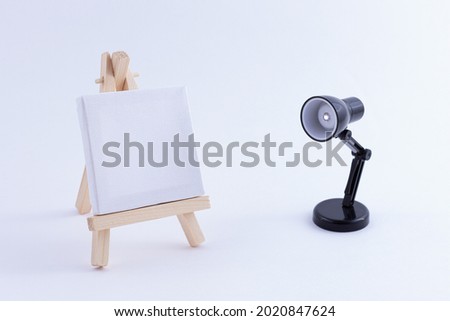 Wooden Easel Miniature with Blank White Square Canvas for Artists and Painters - Mockup. Mini Wooden Stand with Clean Artboard and Small Black Table Lamp on White Background, Copy Space