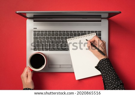 First person top view photo of female hands writing in spiral notebook and holding cup of drink on laptop on isolated red background