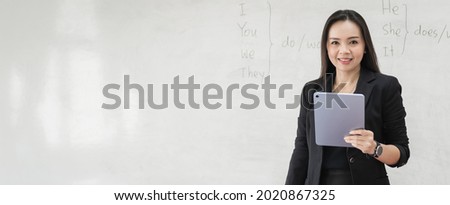 Stock photo portrait of a confident cheerful Asian woman teacher in a black business suit uniform with a digital tablet and laptop to teach modern language in the classroom