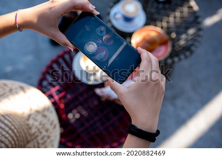 Mobile phone screen detail used as camera to take photos of breakfast at the bar, activities of influencers and bloggers on holiday, heart shape in cappuccino, woman hand holding smartphone
