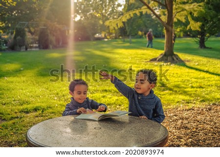 Cute four year old African American twin boys sitting at table in Midwestern park in fall; one boy is looking at picture book while the other is throwing something