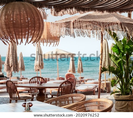 Beach club concept, food corner or cafe, restaurant tables, straw natural chairs, wooden tables, makrome knitted sun umbrellas, white modern sun beds, sea view on background Royalty-Free Stock Photo #2020976252