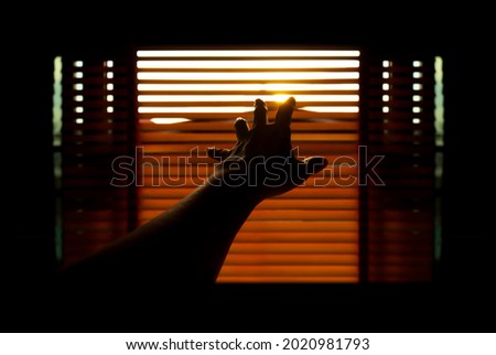 The silhouette of a hand reaching for the light against the blurred background of the blinds in warm tones. The concept of hope and anticipation for new things to come. Royalty-Free Stock Photo #2020981793