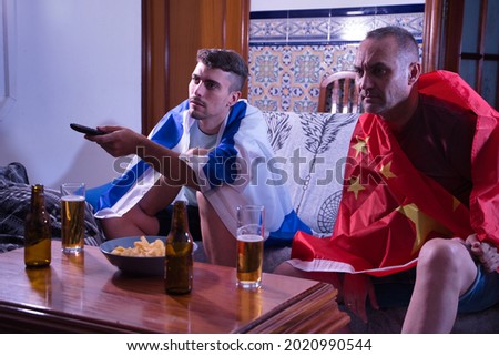 Two men one mature and one young watching sports competitions on television, changing channels with the command with flags of China and Israel.