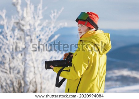 Female photographer in ski equipment with a camera with a huge lens. Winter clothes: red pants and a warm yellow jacket, knitted hat, ski goggles. Selective focus.